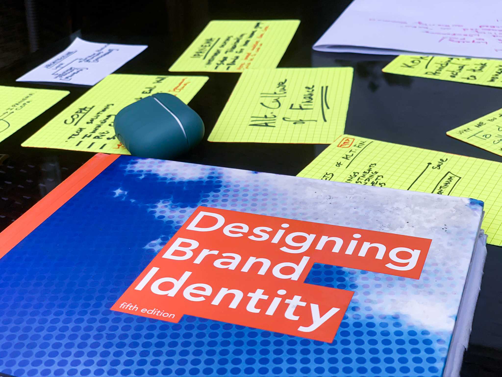 A book called designing brand identity lying on the table with some sticky notes and an airpods pro case